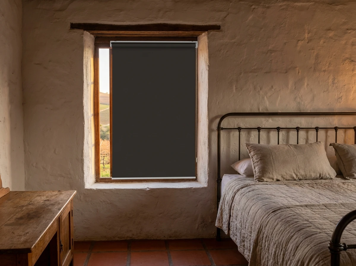 Flat blockout roller blind fully lowered over a small window in a wine-village cottage bedroom near Hermon, Western Cape, vineyard hills visible